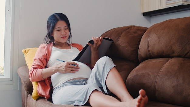 Joven mujer embarazada asiática dibujo bebé en el vientre en el cuaderno. Mamá se siente feliz sonriendo positiva y pacífica mientras cuide al niño acostado en el sofá en la sala de estar en casa.
