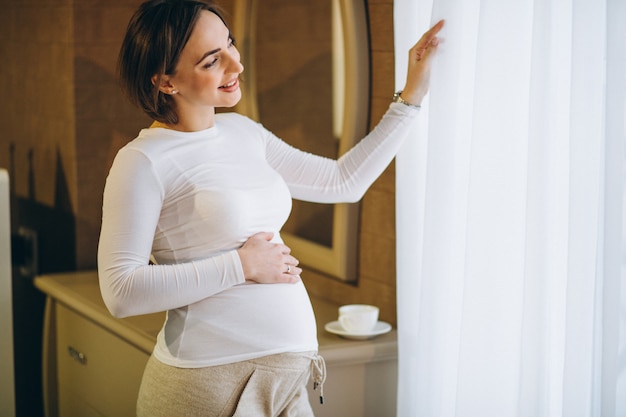 Joven mujer embarazada de pie junto a la ventana y beber café