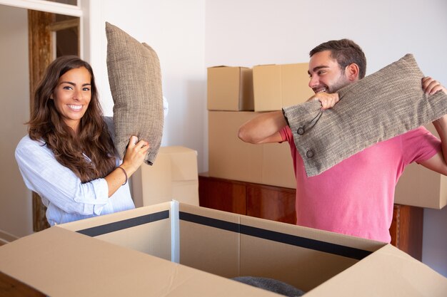 Un joven y una mujer emocionados positivos saliendo de los cojines de la caja de cartón abierta, disfrutando de mover y desempacar cosas