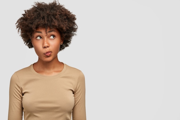 Joven mujer encantadora tiene piel oscura, expresión desorientada e inconsciente, frunce los labios, mira con perplejidad hacia arriba, aislada sobre una pared blanca