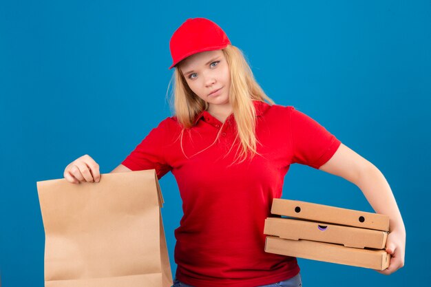 Joven mujer de entrega disgustada vistiendo polo rojo y gorra de pie con paquete de papel y cajas de pizza mirando a cámara sobre fondo azul aislado