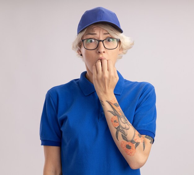 Joven mujer de entrega en uniforme azul y gorra estresada y nerviosa mordiendo las uñas de pie sobre la pared blanca