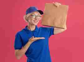 Foto gratuita joven mujer de entrega en uniforme azul y gorra con gafas sosteniendo el paquete de papel sonriendo alegremente presentando con el brazo oh su mano sobre la pared rosa