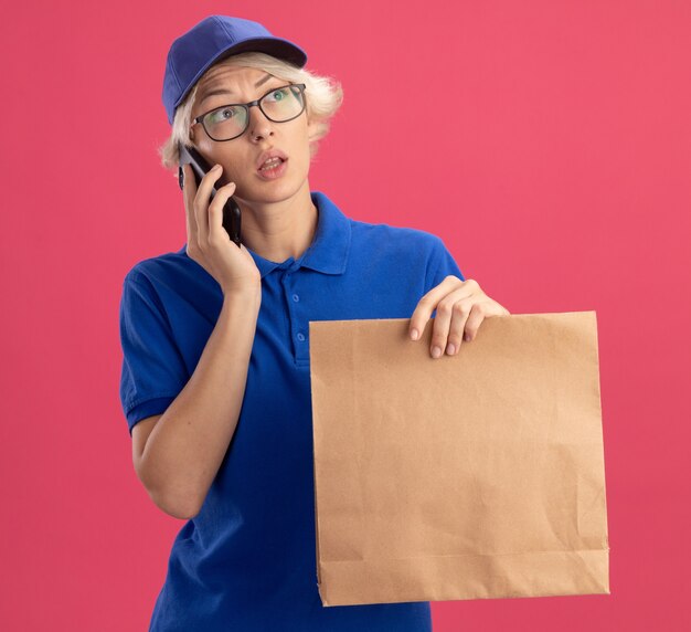 Joven mujer de entrega en uniforme azul y gorra sosteniendo el paquete de papel hablando por teléfono móvil mirando a un lado perplejo sobre la pared rosa