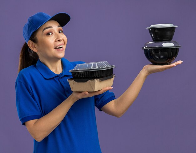 Joven mujer de entrega en uniforme azul y gorra sosteniendo paquetes de alimentos feliz y alegre sonriendo de pie sobre la pared púrpura