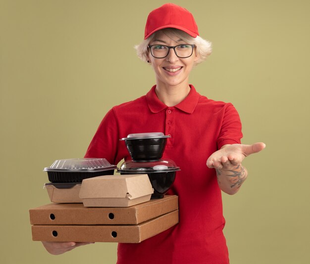 Joven mujer de entrega en uniforme rojo y gorra con gafas sosteniendo cajas de pizza y paquetes de alimentos que ofrecen manos sonriendo de pie amistoso sobre la pared verde