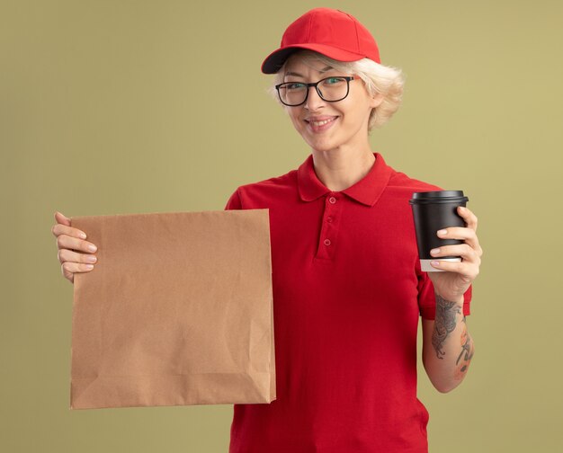 Joven mujer de entrega en uniforme rojo y gorra con gafas sosteniendo el paquete de papel y la taza de papel sonriendo amable de pie sobre la pared verde