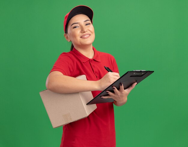 Joven mujer de entrega en uniforme rojo y gorra sosteniendo una caja de cartón con portapapeles escribiendo mirando al frente sonriendo confiado de pie sobre la pared verde