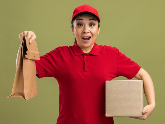 Joven mujer de entrega en uniforme rojo y gorra sosteniendo paquete de papel y caja de cartón feliz y sorprendida de pie sobre la pared verde