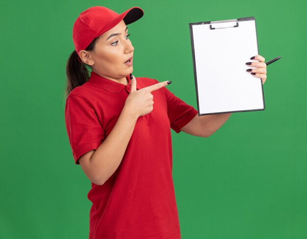 Joven mujer de entrega en uniforme rojo y gorra sosteniendo portapapeles con páginas en blanco apuntando con el dedo índice al portapapeles mirando confundido parado sobre pared verde