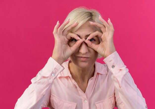 Joven mujer eslava rubia mirando a la cámara y haciendo gesto de mirada con las manos como binoculares aislado sobre fondo rosa