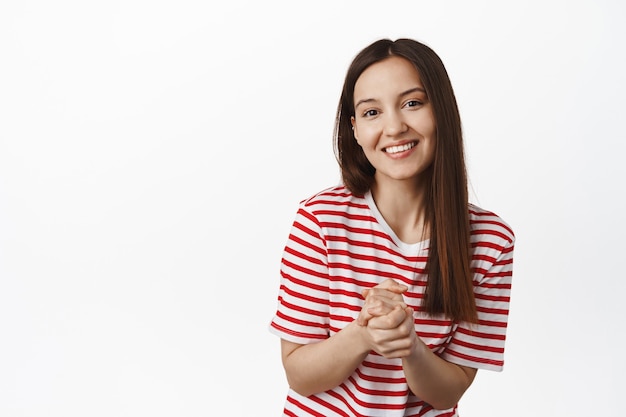 Joven mujer esperanzada aprieta las manos, sonriendo y mirando al frente con expresión amable cortés, esperando algo, pidiendo favor, pared blanca.