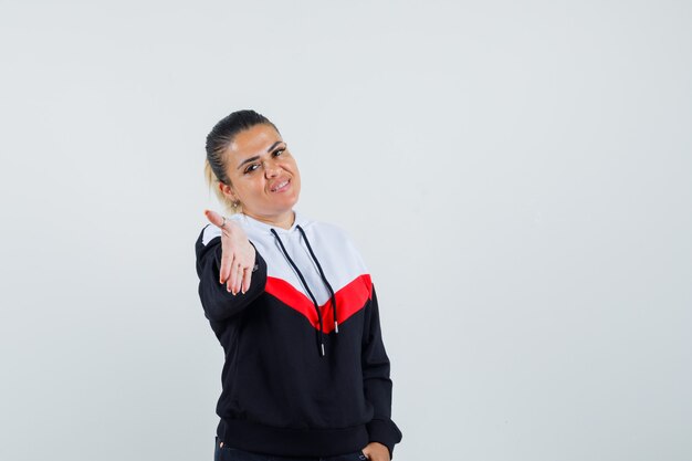 Joven mujer estirando la mano para saludar hacia adelante en colorida sudadera y mirando contento, vista frontal.