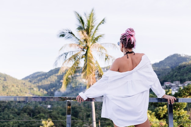 Joven mujer europea en bikini negro de camisa larga blanca con trenzas rosas en la cabeza en palmeras tropicales