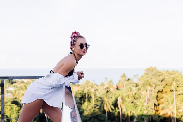 Joven mujer europea en bikini negro de camisa larga blanca con trenzas rosas en la cabeza en palmeras tropicales