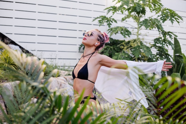 Joven mujer europea en bikini negro de camisa larga blanca con trenzas rosas en la cabeza en palmeras tropicales