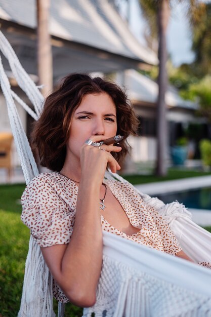 Joven mujer europea fumar cigarro acostado en una hamaca fuera del hotel villa de lujo tropical, luz natural al atardecer