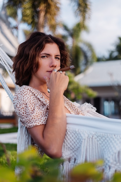 Joven mujer europea fumar cigarro acostado en una hamaca fuera del hotel villa de lujo tropical, luz natural al atardecer