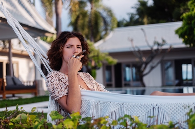 Joven mujer europea fumar cigarro acostado en una hamaca fuera del hotel villa de lujo tropical, luz natural al atardecer