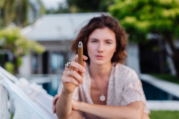 Joven mujer europea fumar cigarro acostado en una hamaca fuera del hotel villa de lujo tropical, luz natural al atardecer