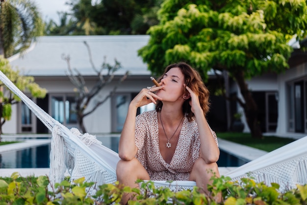 Joven mujer europea fumar cigarro tumbado en una hamaca fuera del hotel villa de lujo tropical