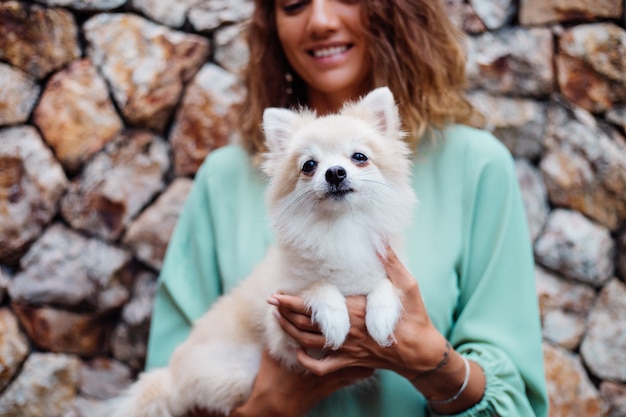 Joven mujer europea en un romántico vestido de verano pulsera sostiene lindo cachorro pomerania esponjoso spitz fuera de villa