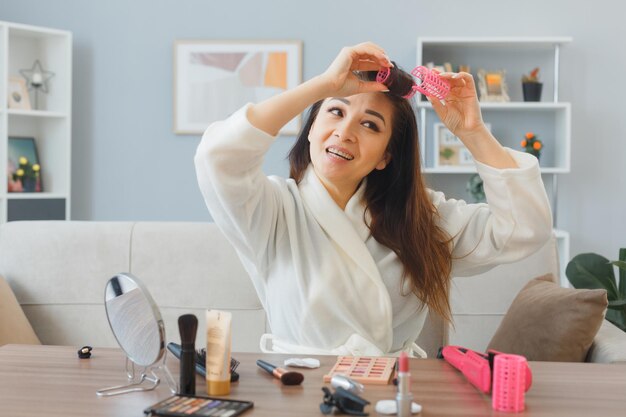 joven mujer feliz con cabello largo y oscuro sentada en el tocador en el interior de la casa aplicando rulos en el cabello haciendo rutina de maquillaje matutino