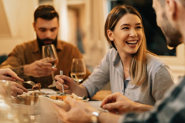 Joven mujer feliz divirtiéndose mientras cena y se comunica con amigos en la mesa del comedor