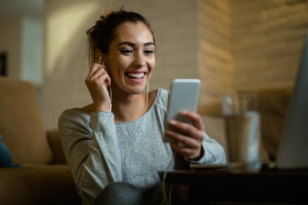 Joven mujer feliz divirtiéndose mientras usa el teléfono móvil y lee mensajes de texto en su sala de estar