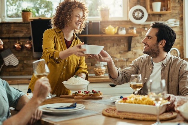 Joven mujer feliz hablando con su amiga mientras trae comida para el almuerzo en la mesa del comedor