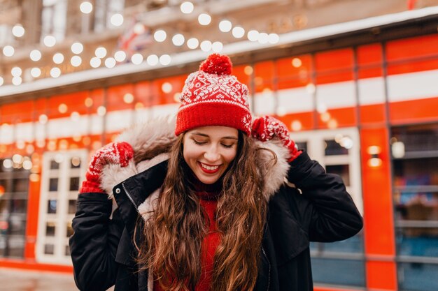 Joven mujer feliz muy sonriente en guantes rojos y gorro de punto con abrigo de invierno caminando en las calles de la ciudad, ropa de abrigo