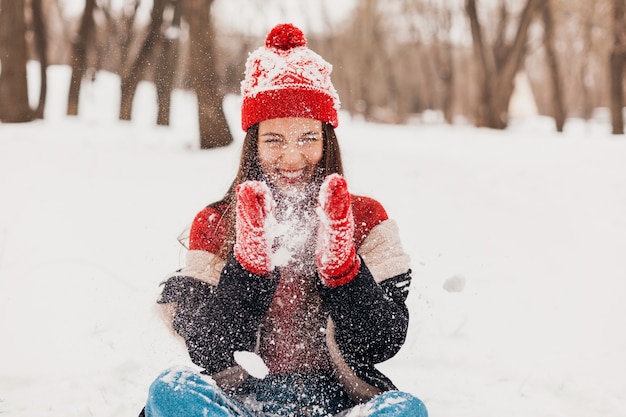 Joven mujer feliz muy sonriente en guantes rojos y gorro de punto con abrigo de invierno sentado sobre la nieve en el parque, ropa de abrigo