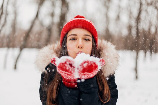 Joven mujer feliz muy sonriente en guantes rojos y gorro de punto vistiendo abrigo de invierno, caminando en el parque, soplando nieve