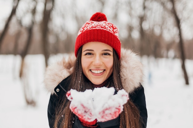 Joven mujer feliz muy sonriente en guantes rojos y gorro de punto vistiendo abrigo de invierno, caminando en el parque, soplando nieve