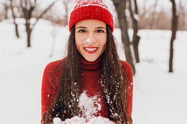 Joven mujer feliz muy sonriente en guantes rojos y sombrero con suéter de punto caminando en el parque en la nieve, ropa de abrigo, divirtiéndose