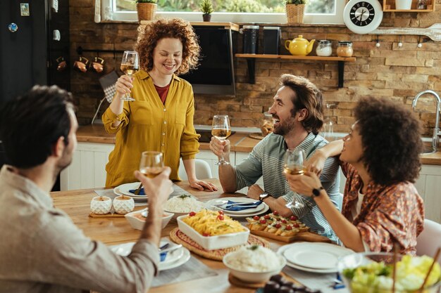 Joven mujer feliz proponiendo un brindis mientras almuerza con sus amigos en casa