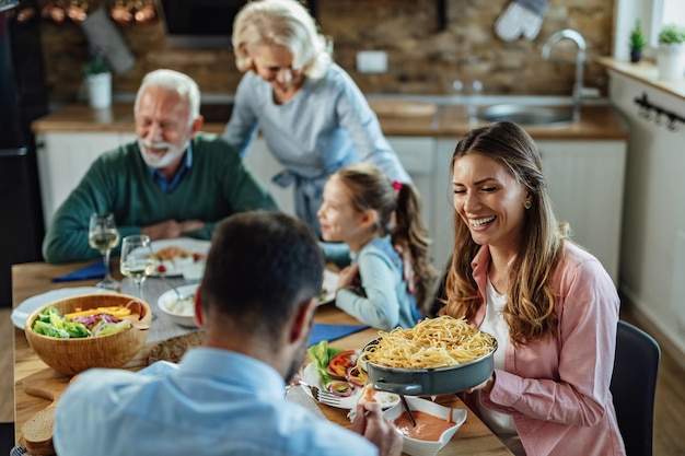 Joven mujer feliz riéndose mientras le pasa comida a su marido y disfruta del almuerzo familiar en la mesa del comedor.