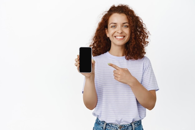 Foto gratuita joven mujer feliz señalando con el dedo la pantalla del teléfono móvil y sonriendo complacida, recomendando una aplicación, una aplicación útil, una buena interfaz, fondo blanco