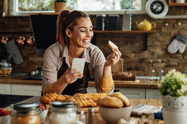 Joven mujer feliz sosteniendo una rebanada de pan mientras usa un teléfono inteligente y prepara comida en la cocina