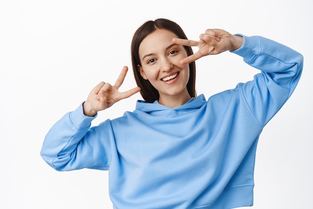 Joven mujer feliz en sudadera con capucha azul, mostrando signos v de paz, gesto disco cerca de los ojos, bailando y sonriendo alegre, de pie sobre una pared blanca