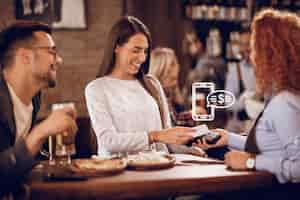 Foto gratuita joven mujer feliz usando un teléfono inteligente mientras paga una factura a una camarera en un bar