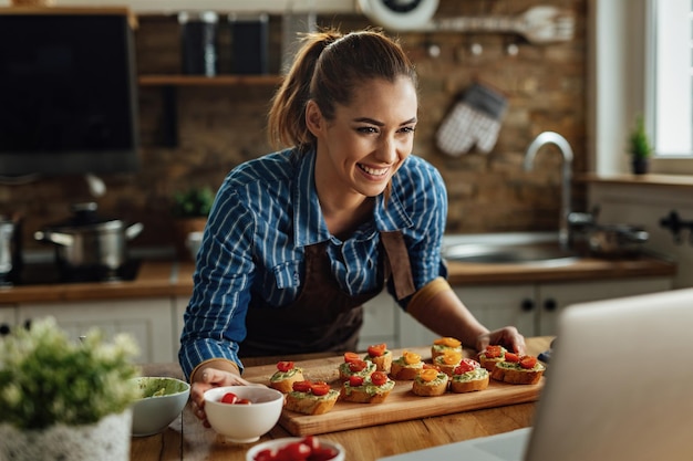 Joven mujer feliz vlogueando sobre una laptop mientras prepara bruschetta con aguacate y tomate cherry en la cocina