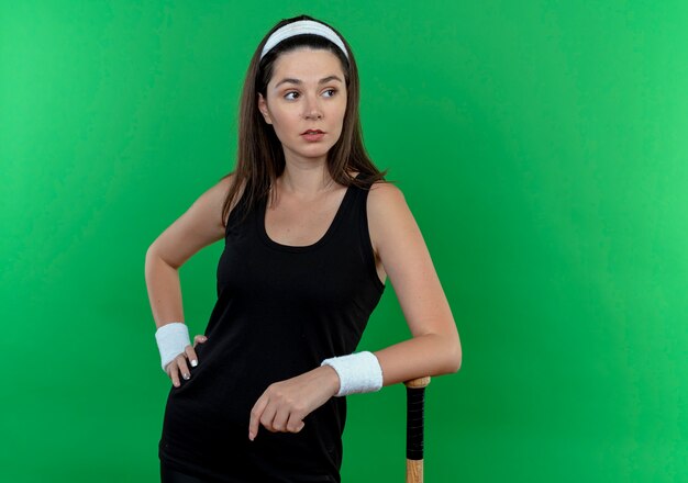 Joven mujer fitness en diadema apoyando su mano en el bate de béisbol mirando a un lado confiados de pie sobre la pared verde