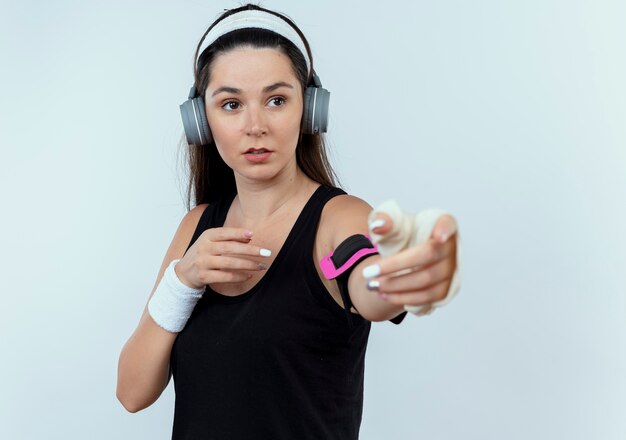 Joven mujer fitness en diadema con auriculares y brazalete de smartphone mirando a un lado con los brazos extendidos de pie sobre la pared blanca
