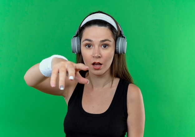 Joven mujer fitness en diadema con auriculares con expresión confusa con el brazo extendido de pie sobre la pared verde