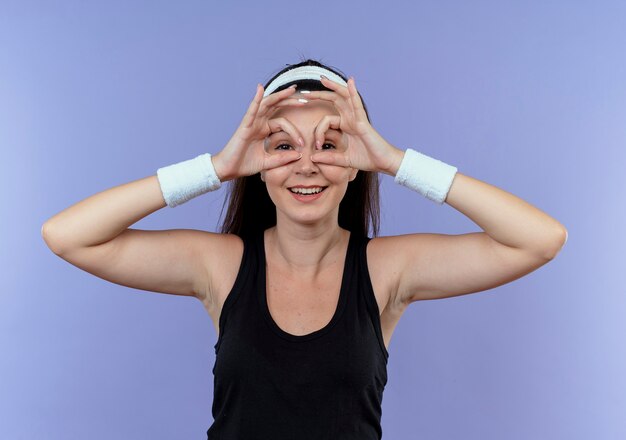 Joven mujer fitness en diadema haciendo gesto binocular con los dedos a través de los dedos sonriendo de pie sobre la pared azul