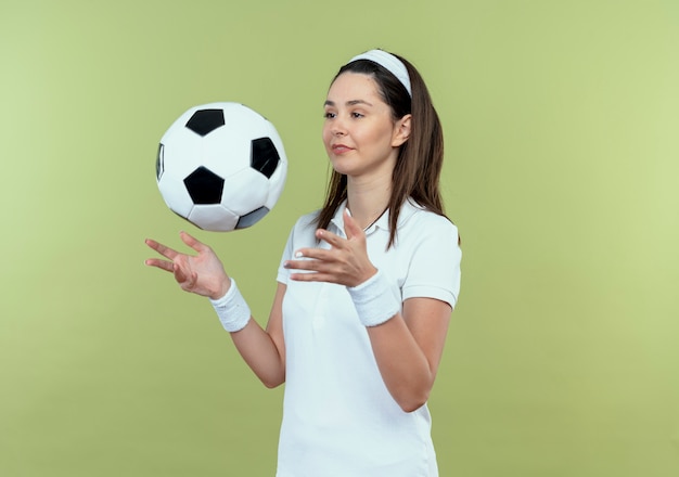 Joven mujer fitness en diadema lanzando una pelota de fútbol sonriendo confiados de pie sobre la pared de luz