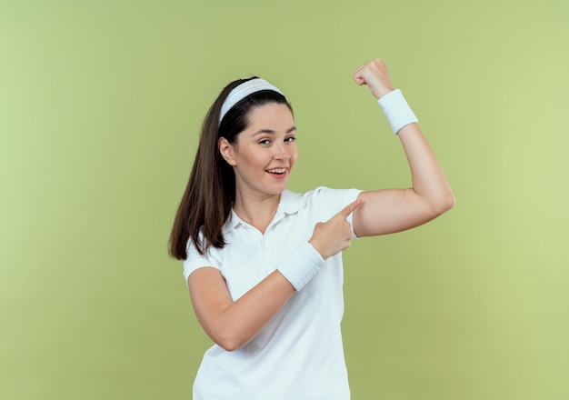 Joven mujer fitness en diadema levantando puño mostrando bíceps sonriendo confiados de pie sobre la pared de luz