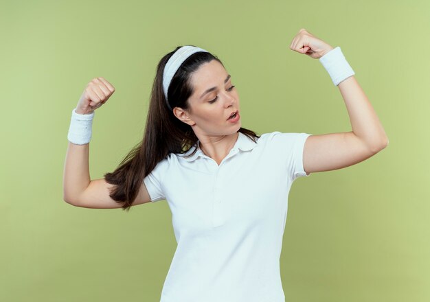 Joven mujer fitness en diadema levantando puños mostrando bíceps mirando confiados de pie sobre la pared de luz