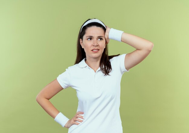 Joven mujer fitness en diadema mirando a un lado perplejo con la mano en la cabeza de pie sobre la pared de luz